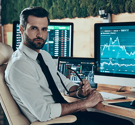 Professional financial trader sitting at a desk with multiple market data monitors looking directly at the camera.